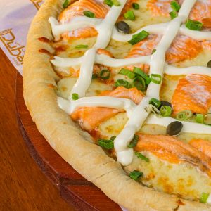 A close-up of a gourmet salmon pizza with cream cheese, green onions, and capers on a wooden surface.
