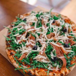 Close-up of a delicious arugula and prosciutto pizza with cheese and olives on a wooden table.
